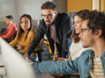 Erwachsene sitzen vor Rechnern in einem Seminarraum. Der Dozent erklärt etwas.